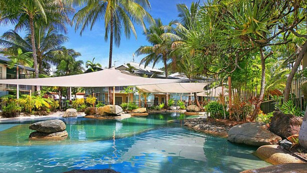 A pool area with clear water, surrounded by rocks and palm trees, and a covered seating area with umbrellas. - Wallaby Hideaway (Trinity Beach)
