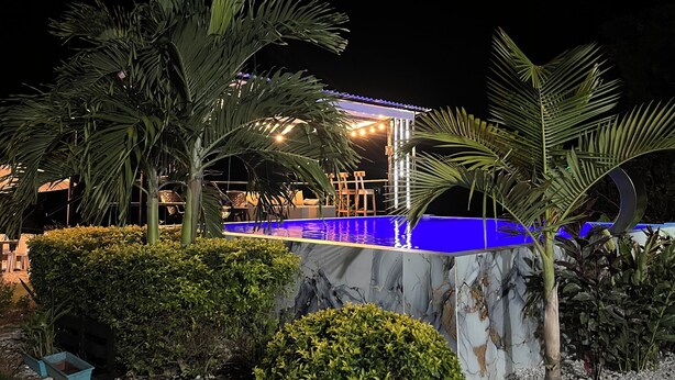 A nighttime pool area with a blue pool, palm trees, and a covered seating area with string lights.