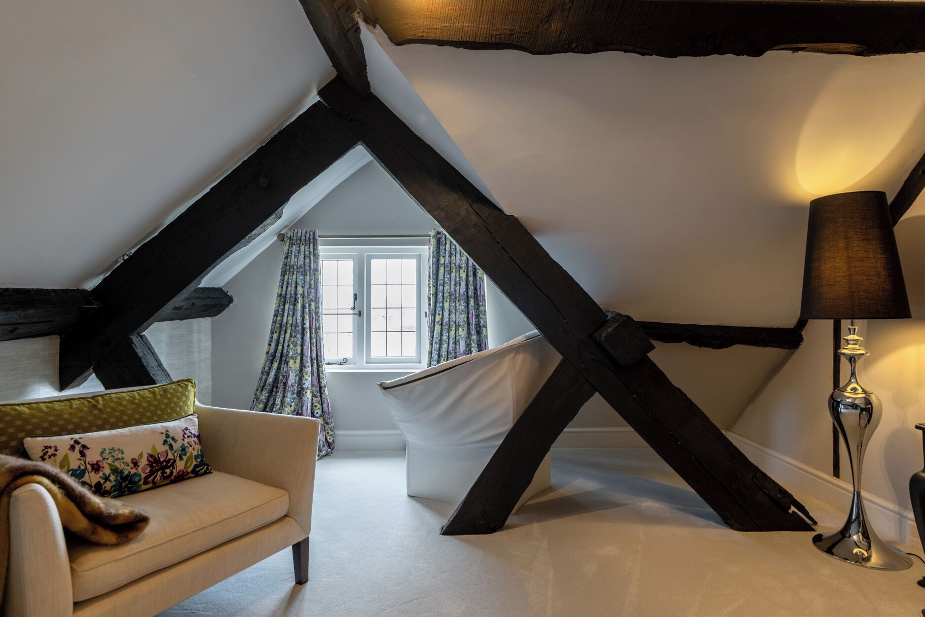 Une chambre confortable située dans les combles, avec des poutres apparentes, un canapé blanc, une lampe et une fenêtre avec des rideaux.