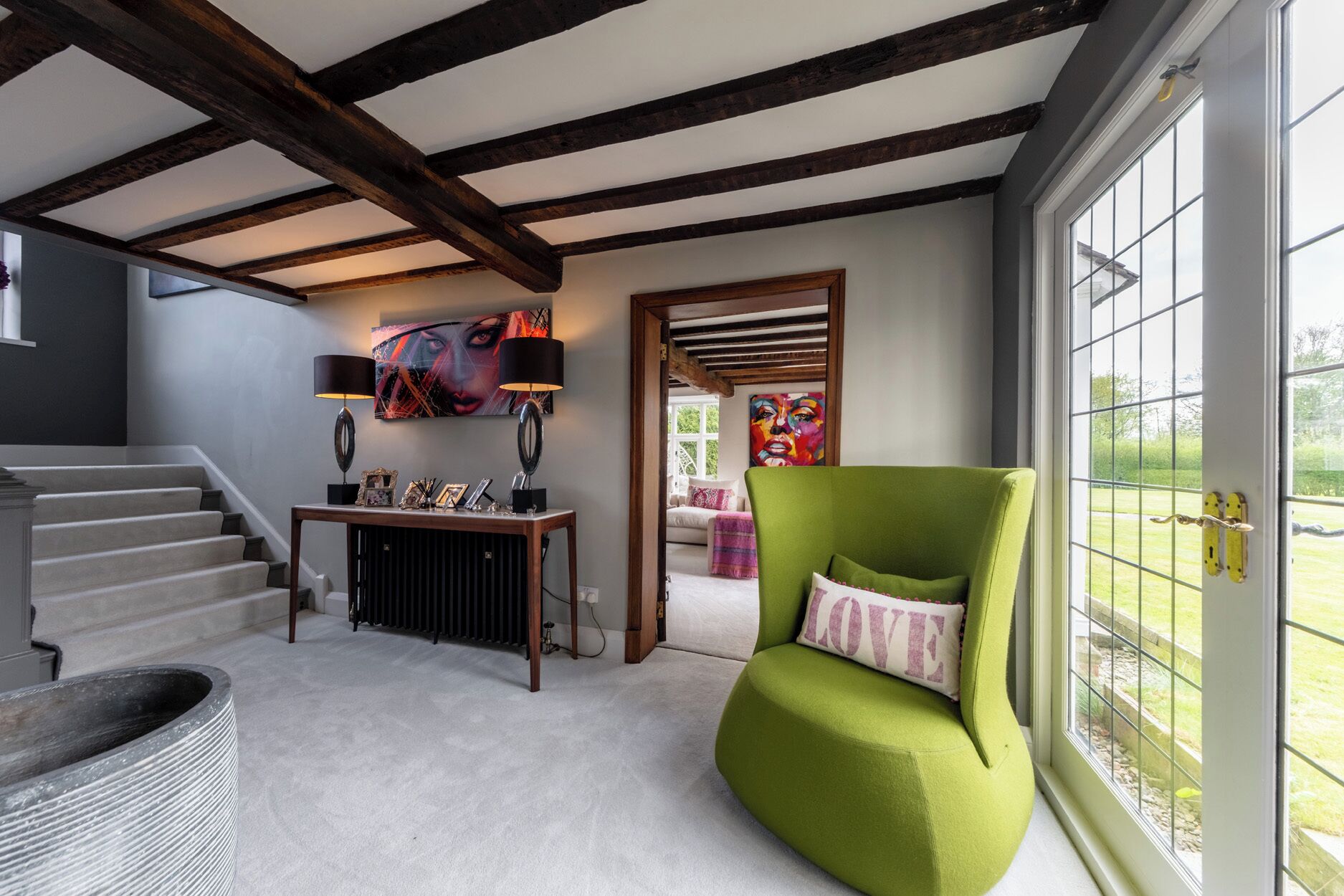 A room with a staircase, a green armchair, a console table with decorative items, and a large window with a view of the outdoors.