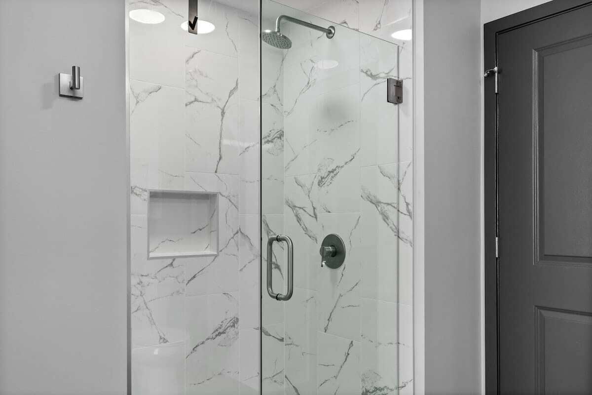 Une douche fermée par des parois de verre, avec un mur décoré de motifs en marbre, une petite étagère et une porte située à droite.