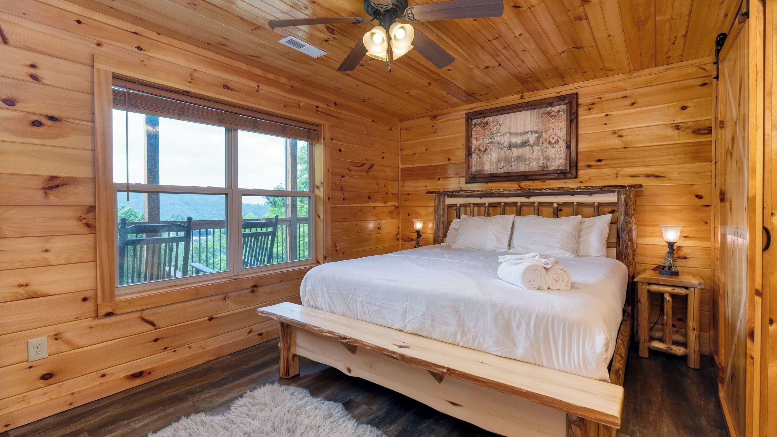A wooden cabin room with a bed, a bench, a ceiling fan, a window, and a painting.