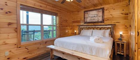 A wooden cabin room with a bed, a bench, a ceiling fan, a window, and a painting.