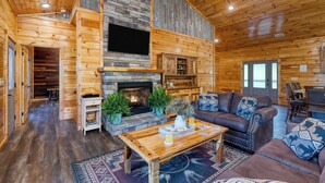 A log cabin interior with a stone fireplace, wooden walls, a sofa, and a dining area. - Evergreen Echo by Avantstay Cabin w/ Mountain Views, Theater, Game Room, and Hot Tub (Gatlinburg)