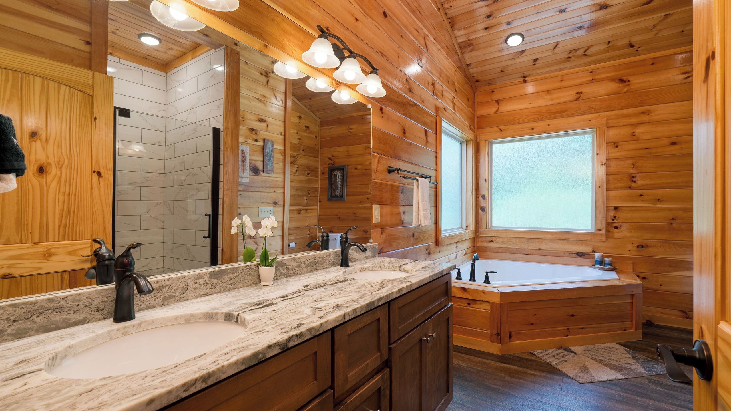 A wooden cabin bathroom with a large mirror, two sinks, a bathtub, and a window.