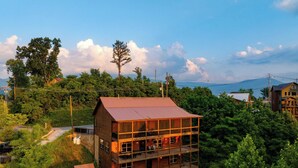 A multi-story wooden building with balconies, surrounded by lush greenery and a mountainous backdrop. - Evergreen Echo by Avantstay Cabin w/ Mountain Views, Theater, Game Room, and Hot Tub (Gatlinburg)