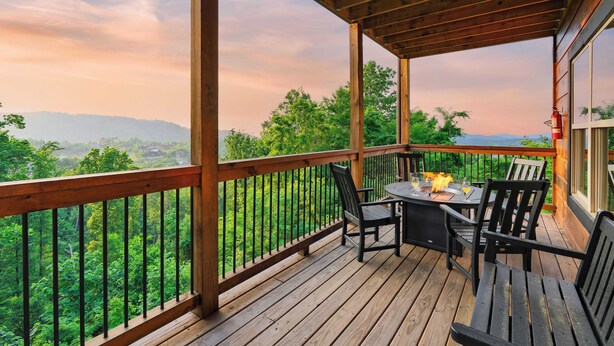 A wooden deck with a table and chairs, overlooking a lush green landscape. - Evergreen Echo by Avantstay Cabin w/ Mountain Views, Theater, Game Room, and Hot Tub (Gatlinburg)