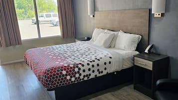 A hotel room with a large bed, a nightstand, a desk, and a chair. There is a window with curtains and a view of the outside.