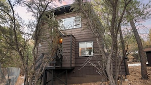 Una cabaña de madera de dos pisos con una escalera que lleva al nivel superior, rodeada de árboles y un área cercada.