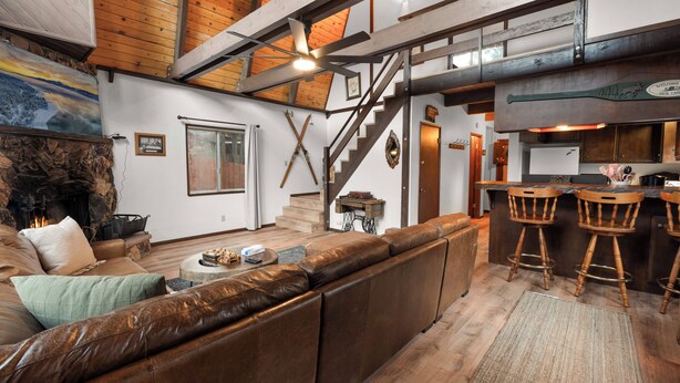 Amplia sala de estar con sofá de cuero, piso de madera, chimenea y una cocina con taburetes de bar.