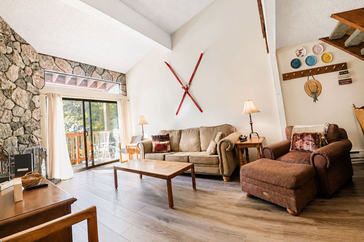 A spacious living room with a stone wall, a large window, a wooden coffee table, and a red X-shaped decoration on the wall.