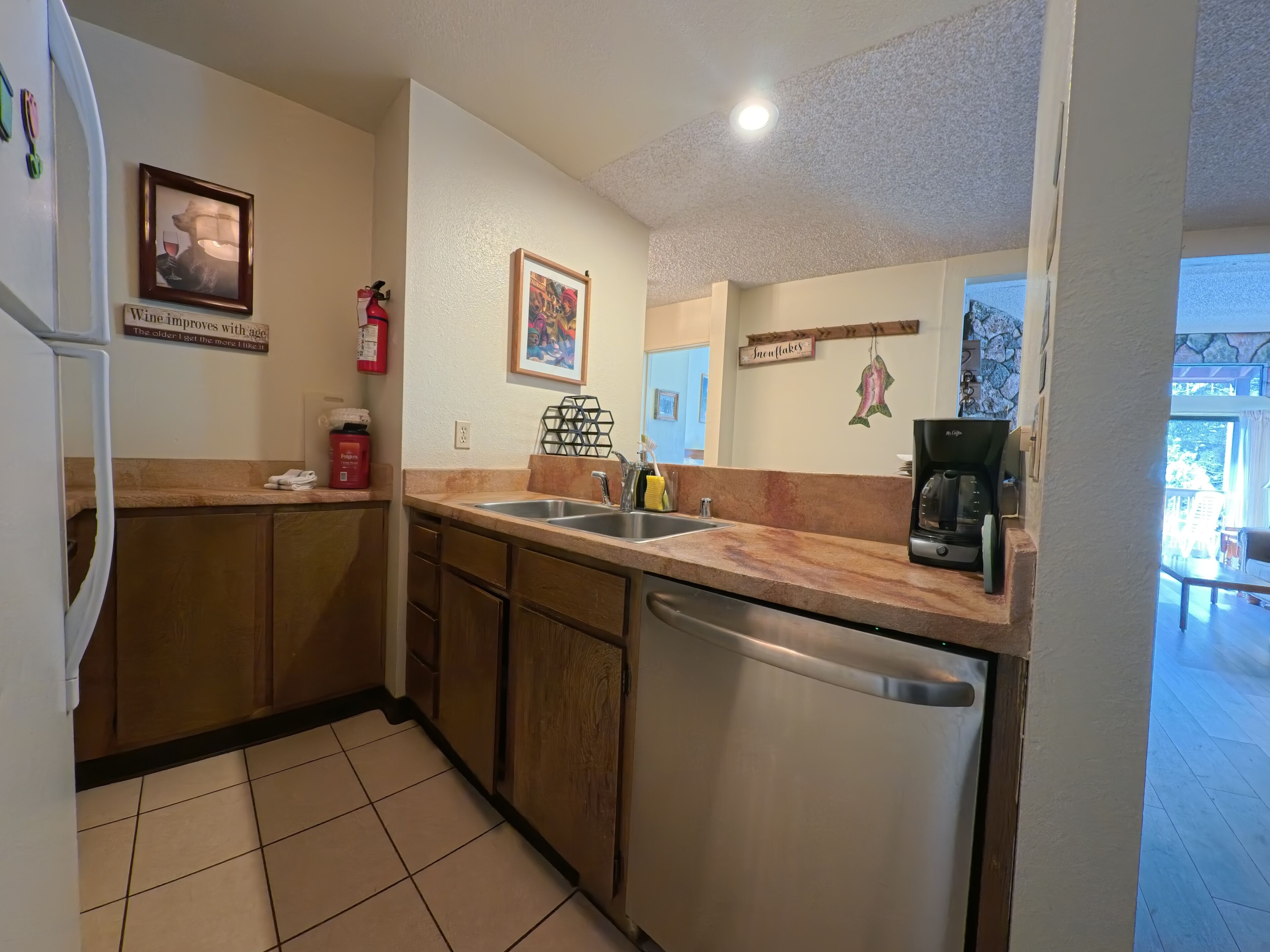 A kitchen with wooden cabinets, a dishwasher, a coffee maker, and a sink. There are framed pictures on the wall and a fire extinguisher.