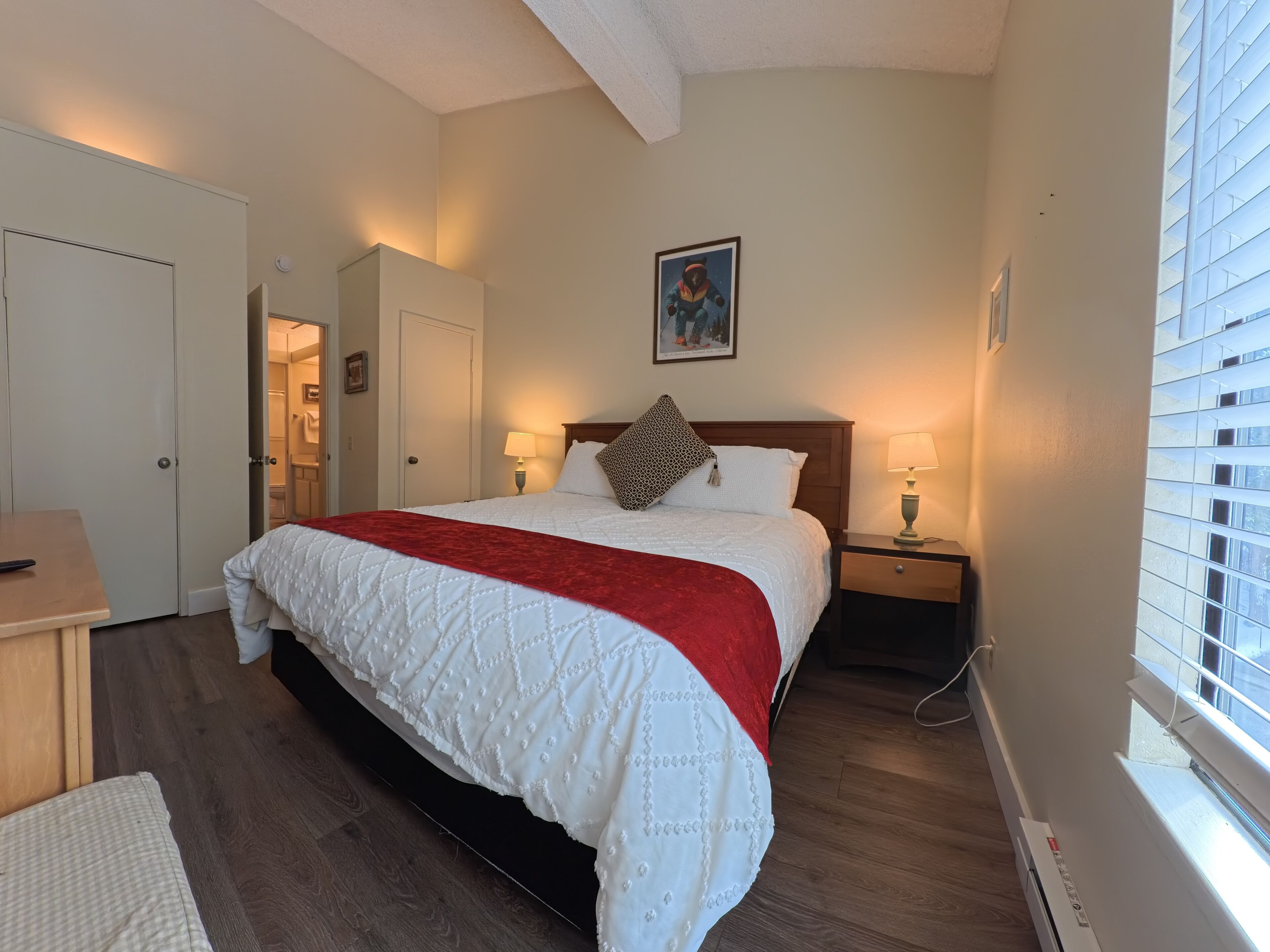 Une chambre à coucher avec un grand lit, une table de chevet avec une lampe, un bureau en bois et une fenêtre avec des stores.