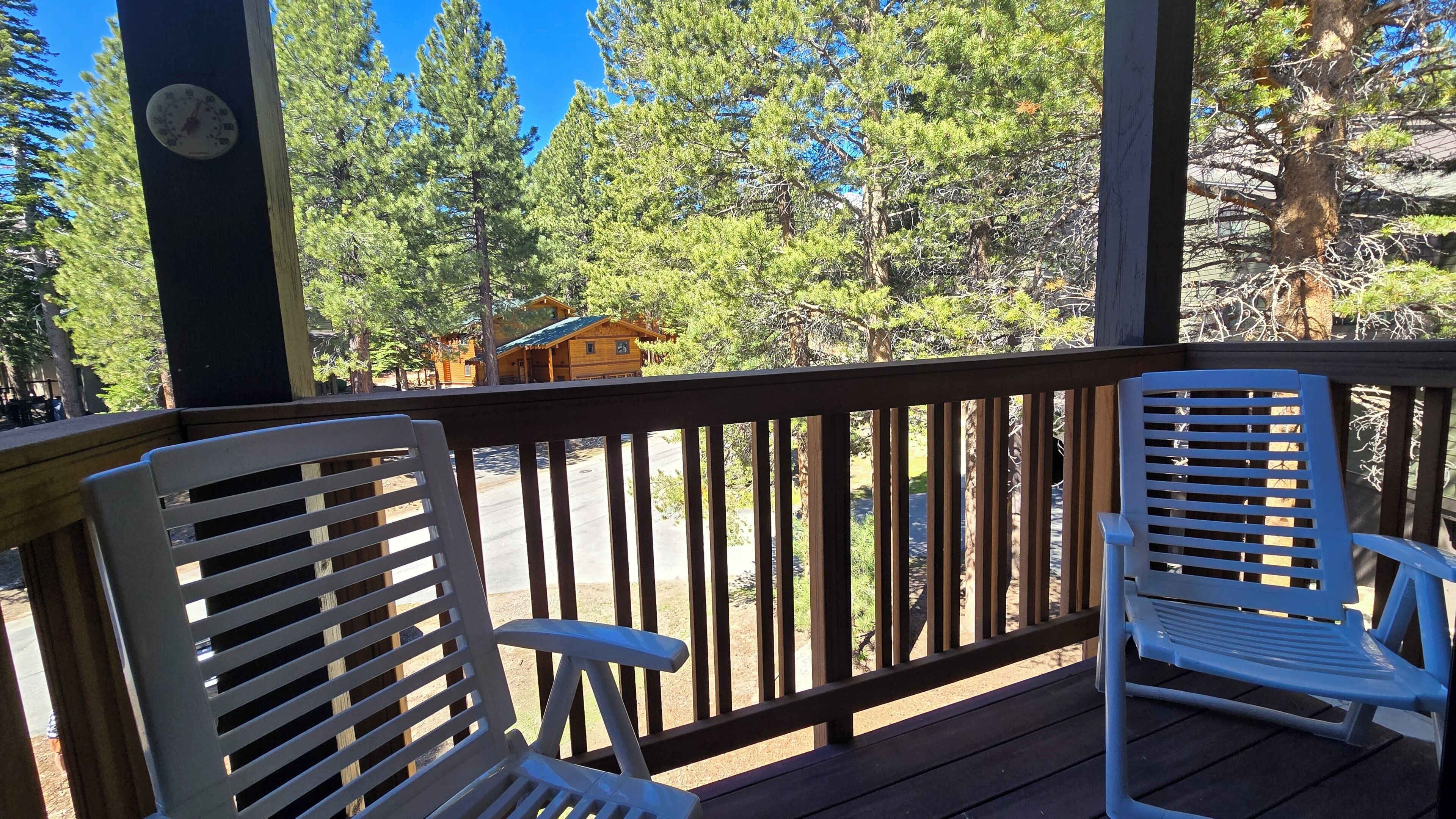 A wooden deck with two white chairs, a wooden railing, and a view of a cabin surrounded by trees.