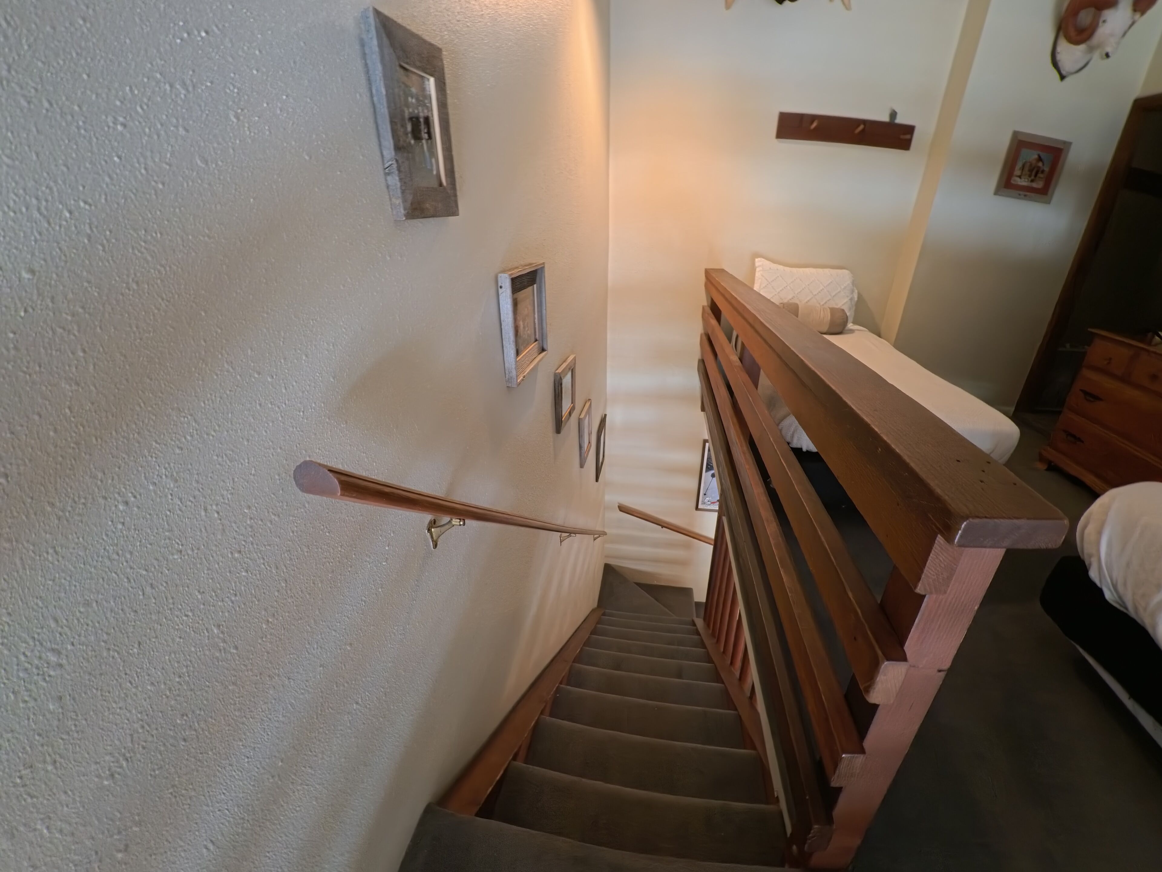 A staircase with wooden handrails, a bed with a white mattress and a quilted pillow, and a wooden dresser.