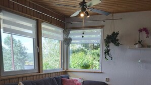 En hyggelig stue med en mørk sofa, et træbord og en loftsvifte. Store vinduer giver naturligt lys og udsigt til grønne omgivelser.