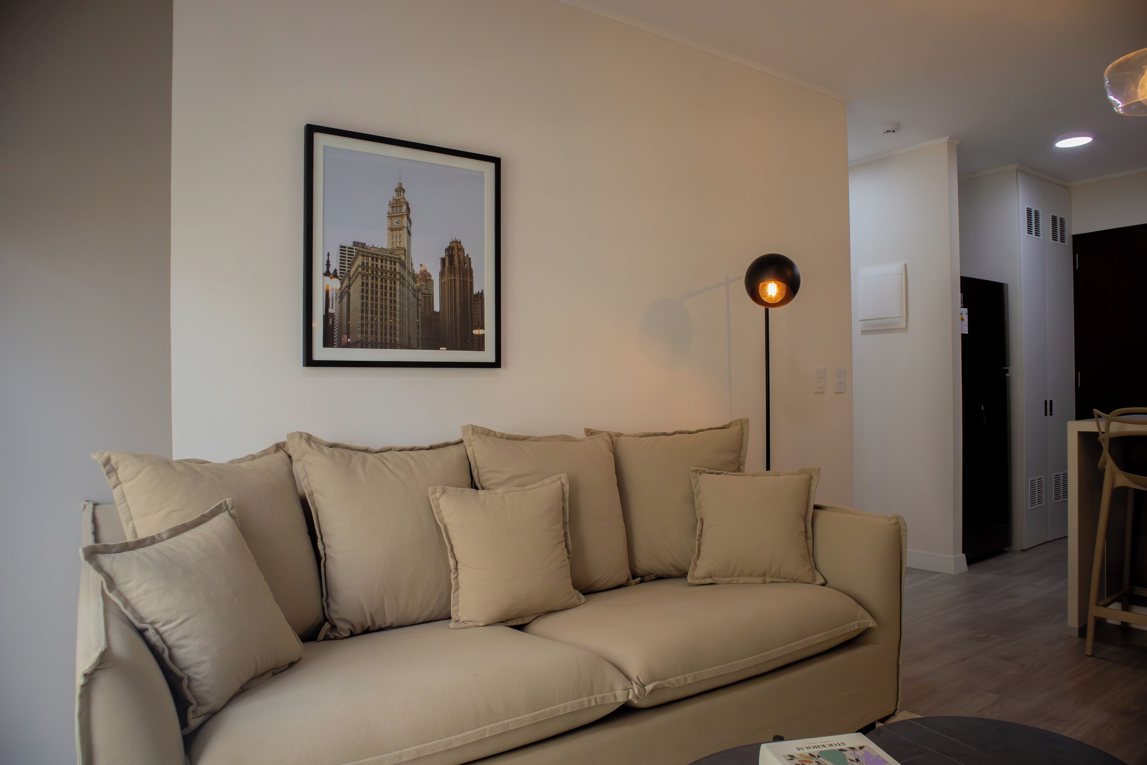 A beige sofa with multiple cushions, a framed cityscape picture on the wall, a floor lamp, and a dining area with a chair and table.
