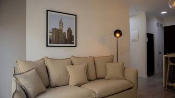 A beige sofa with multiple cushions, a framed cityscape picture on the wall, a floor lamp, and a dining area with a chair and table.
