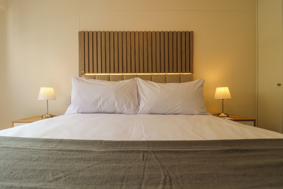 Ein ordentlich bezogenes Bett mit weißen Leinen und einer grauen Tagesdecke, an beiden Seiten mit Nachttischen und Lampen, in einem Zimmer mit einem Holzkopfteil und neutralen Wänden.