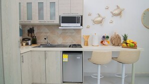 Een compacte keuken met een magnetron, spoelbak en een kleine koelkast. Op het aanrecht staan twee barkrukken en een fruitmand.