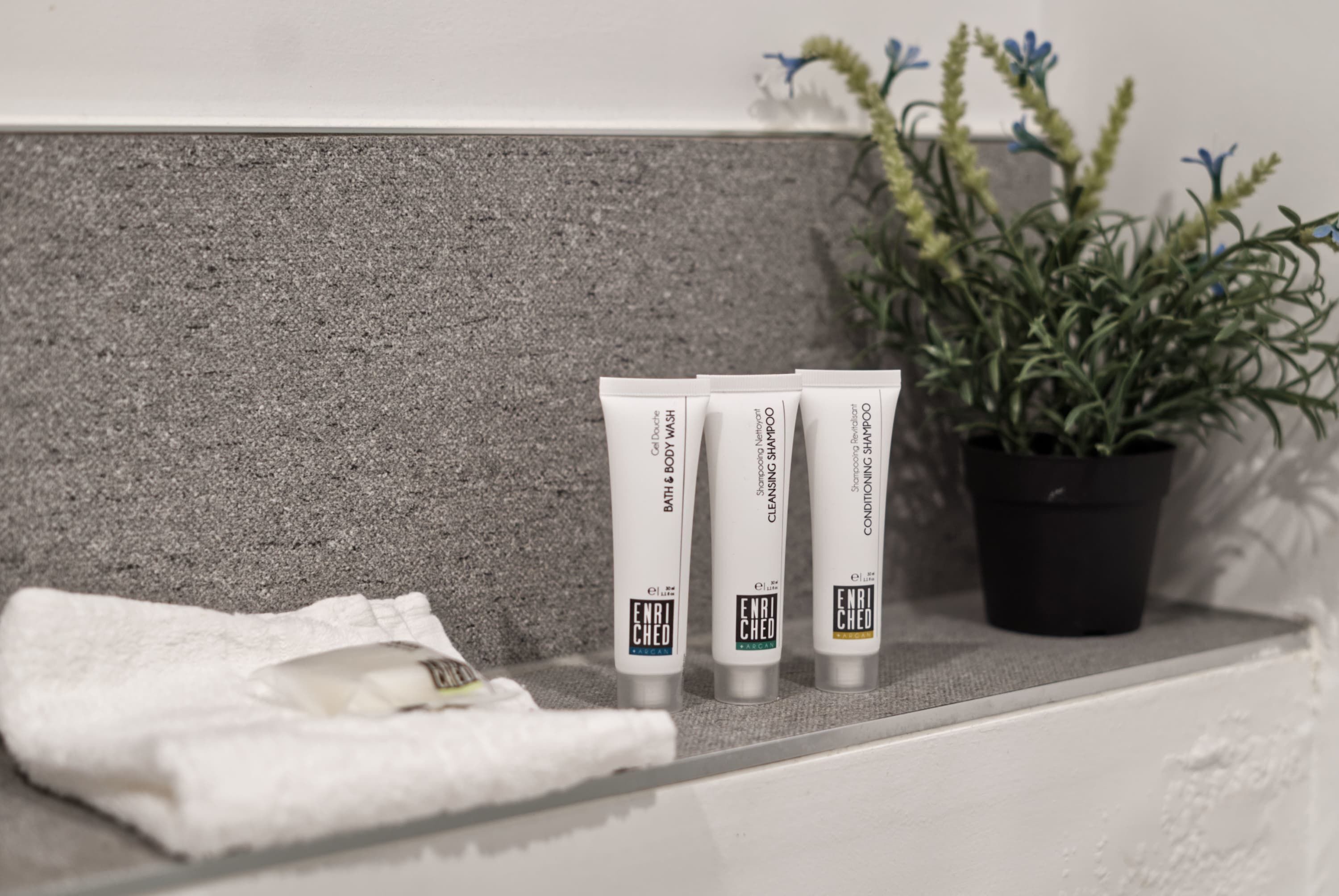 A bathroom shelf with three tubes of Enriched Bath & Body products, a white towel, and a potted plant.
