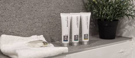 A bathroom shelf with three tubes of Enriched Bath & Body products, a white towel, and a potted plant.