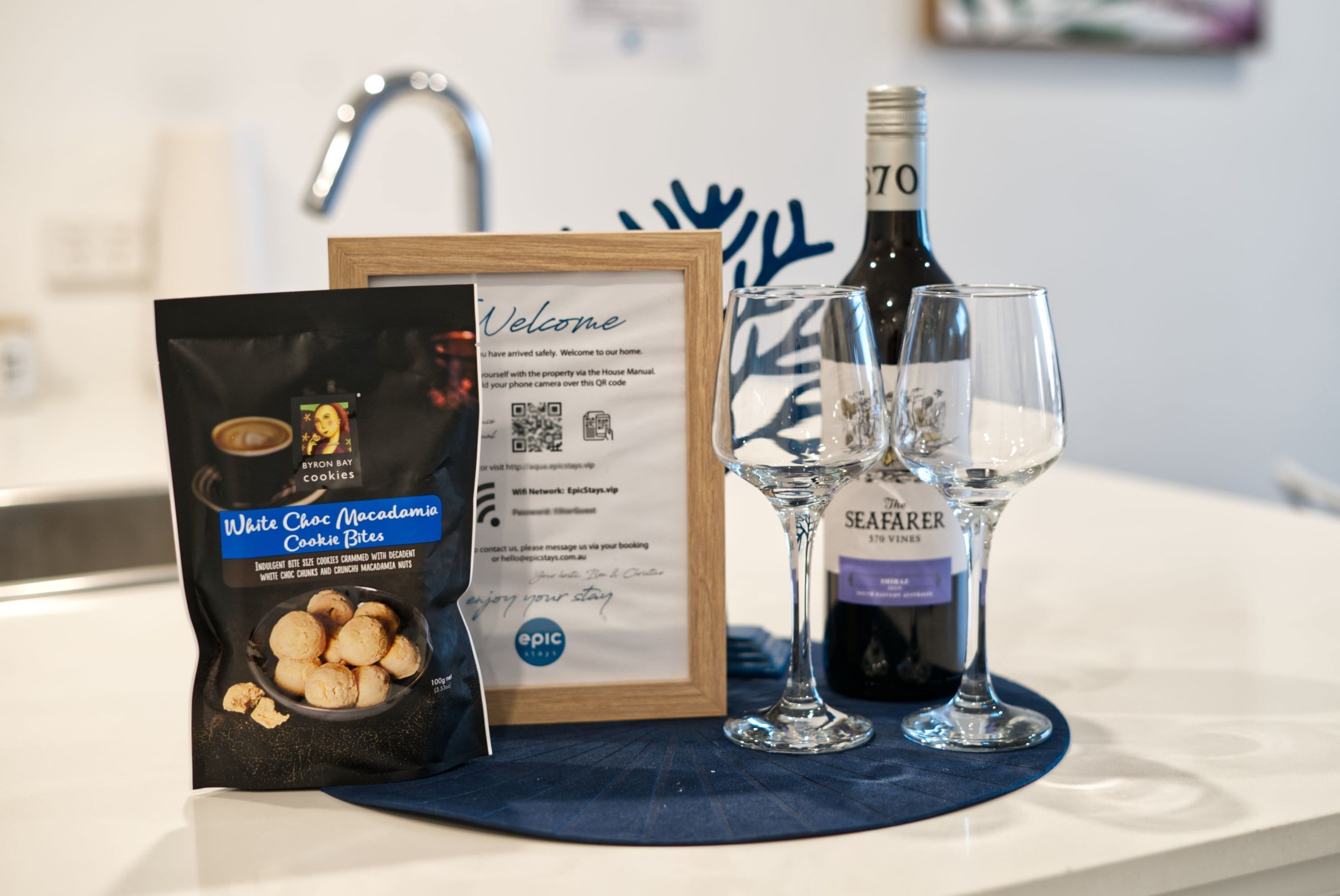 A welcome sign, a bottle of Seafarer wine, two wine glasses, and a bag of White Choc Macadamia Cookie Bites on a counter.