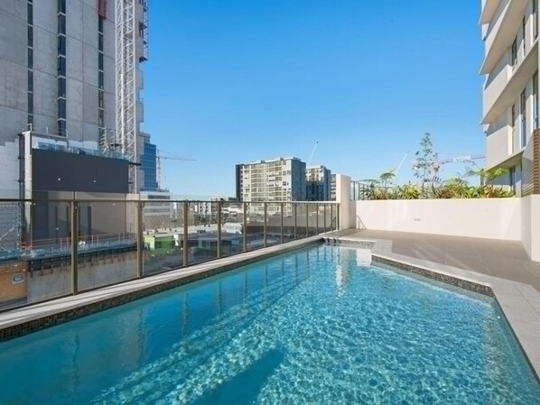 A rooftop swimming pool with clear blue water, surrounded by modern buildings and a clear sky.