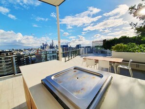 A rooftop terrace with a view of the city skyline, a table, chairs, and a grill.
