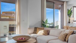 A modern living room with a beige sectional sofa, a small round table with a woven basket, and a large window offering a city view.