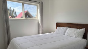 A bed with a white quilted bedspread and two pillows, a wooden headboard, and a window with curtains. - Aspen Cabin at Candle Lake Golf Resort (Candle Lake)