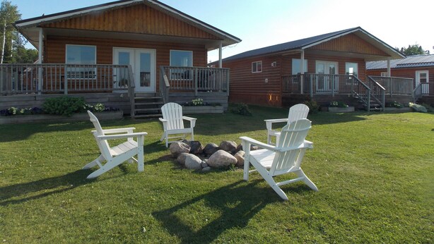 Two wooden cabins with porches, a small fire pit, and white Adirondack chairs in a grassy yard. - Aspen Cabin at Candle Lake Golf Resort (Candle Lake)
