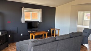 A modern living room with a grey sofa, a wooden coffee table, a flat-screen TV, and a window with curtains. - Candle Lake Golf Course - Spruce Cabin (Candle Lake)