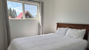 A bed with a white quilted bedspread and two pillows, a wooden headboard, and a window with curtains. - Candle Lake Golf Course - Spruce Cabin (Candle Lake)