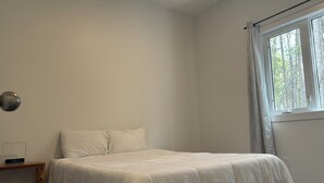 Une chambre simple et épurée, avec un lit, une table de chevet dotée d’une horloge et une fenêtre avec des rideaux.