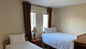 Un dormitorio con dos camas individuales, una mesita de noche con lámpara y una ventana con cortinas.