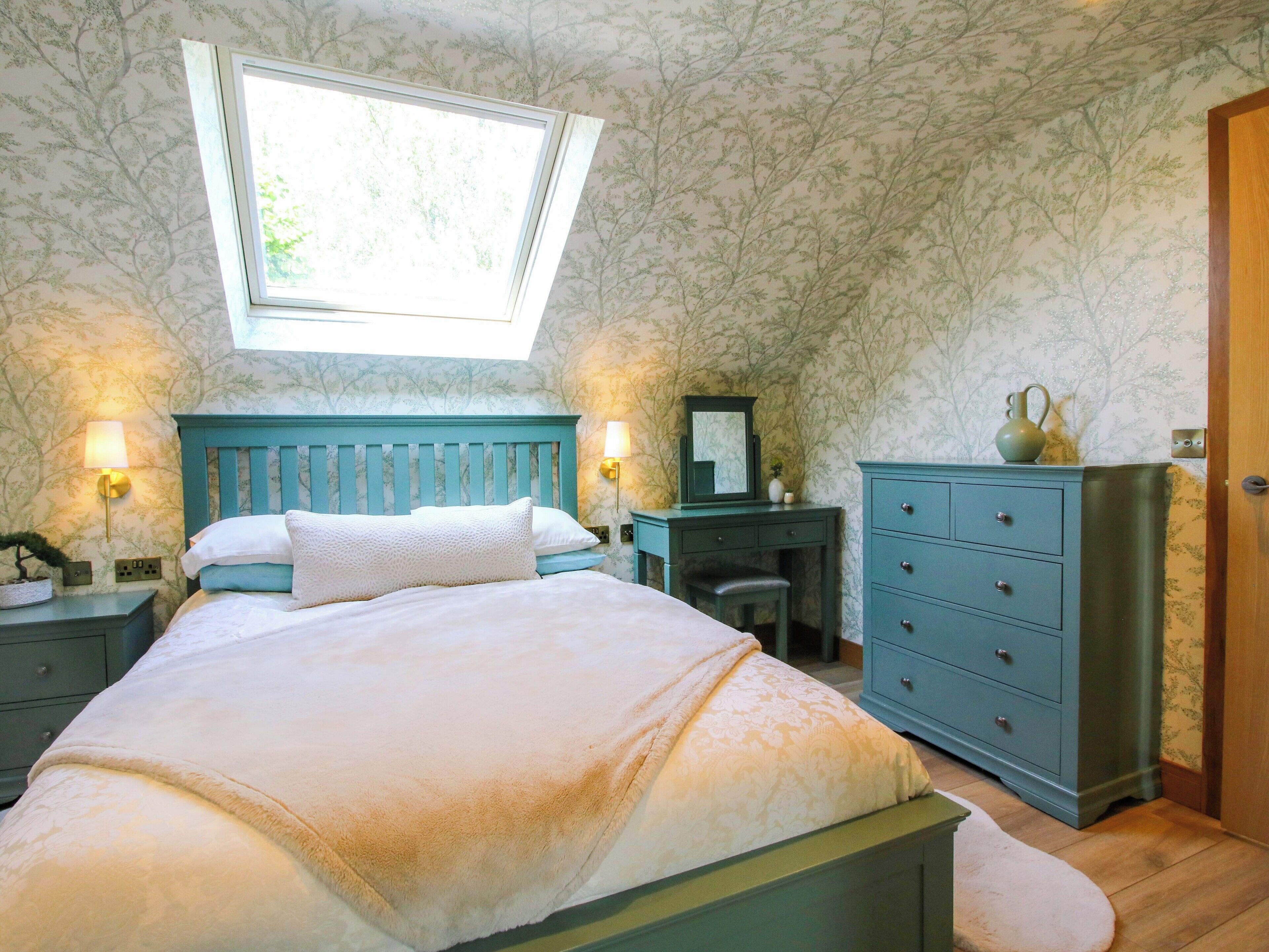 Een slaapkamer met dakraam, een bed met witte lakens, een nachtkastje met lamp, een kaptafel en een spiegel.