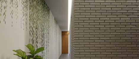 A corridor with a brick wall on the right and a wall with a green leafy pattern on the left. There is a potted plant in the center.