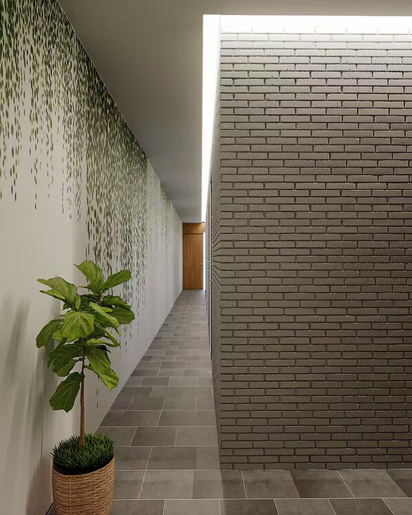 A corridor with a brick wall on the right and a wall with a green leafy pattern on the left. There is a potted plant in the center.