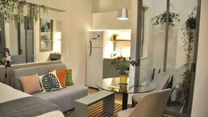 Ein modernes Wohnzimmer mit einer Couch, einem Glastisch und einem Sessel. Durch eine Glasscheibe ist eine Küchenzeile mit Kühlschrank und Spüle erkennbar.