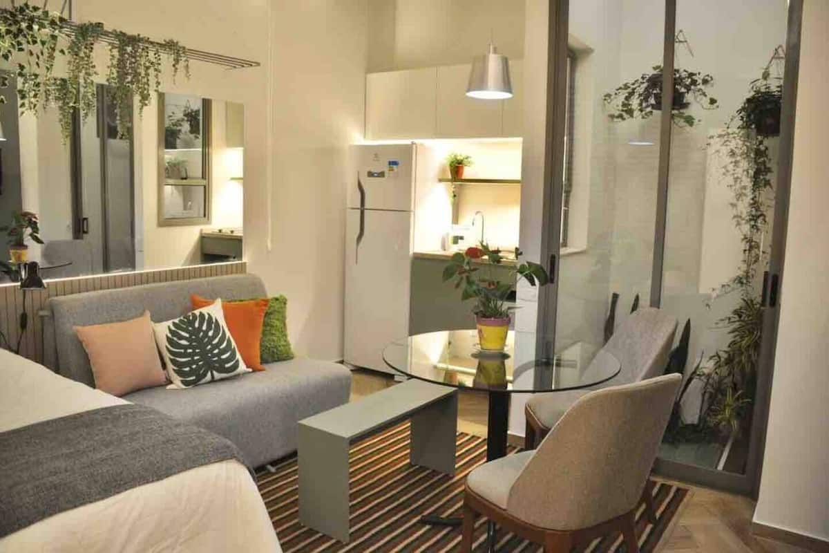 Una sala moderna con un sofá, una mesa de centro de vidrio y una silla. Se observa un área de cocina con un refrigerador y un lavamanos separados por una pared de vidrio.