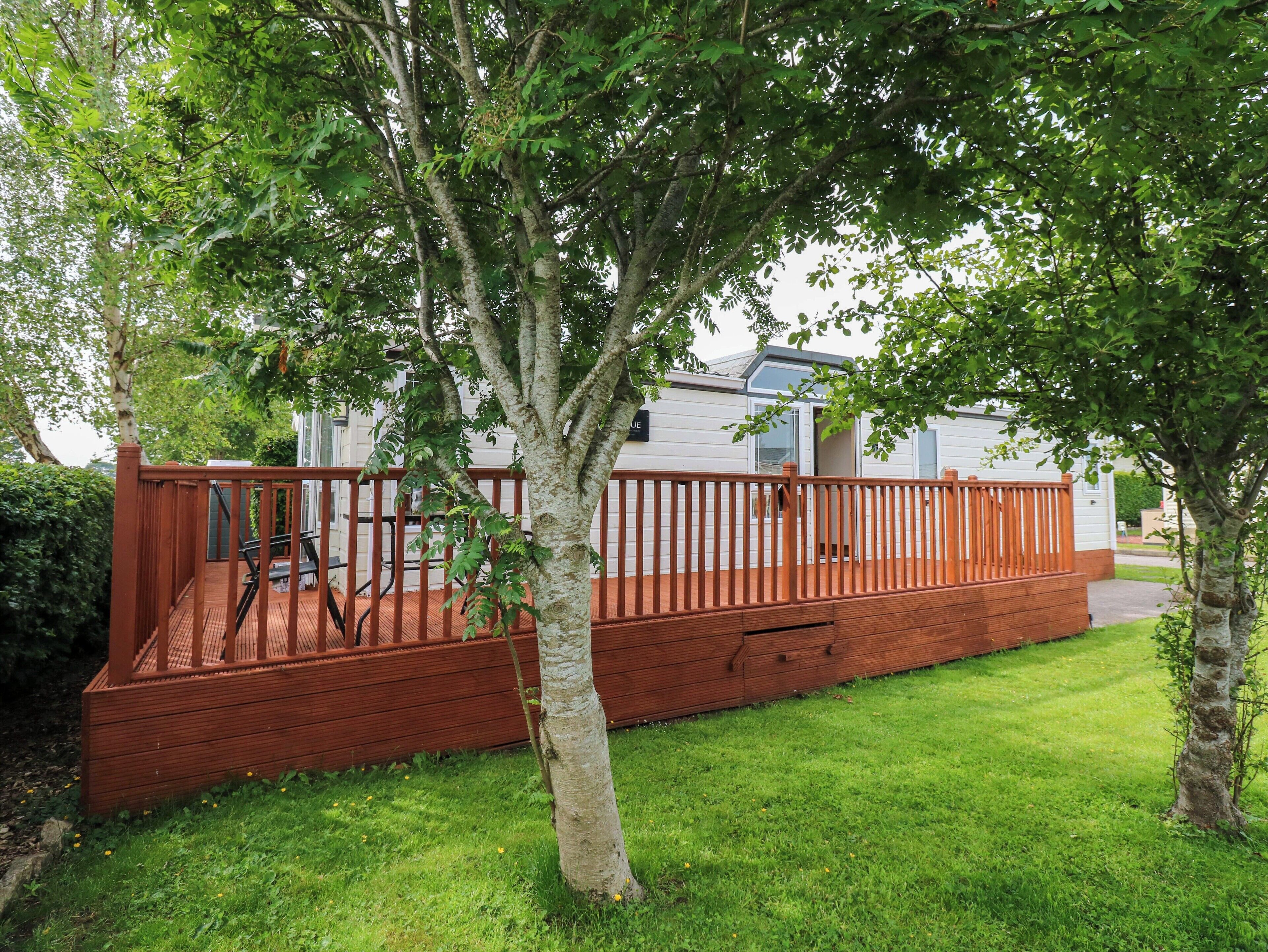 Une terrasse en bois avec un banc intégré, entourée d’un bâtiment blanc doté d’une porte vitrée, située dans un espace verdoyant avec des arbres.