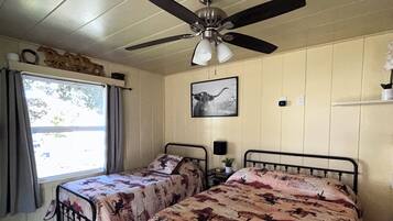 Une chambre avec deux lits, un ventilateur de plafond, une fenêtre avec des rideaux et un tableau accroché au mur.