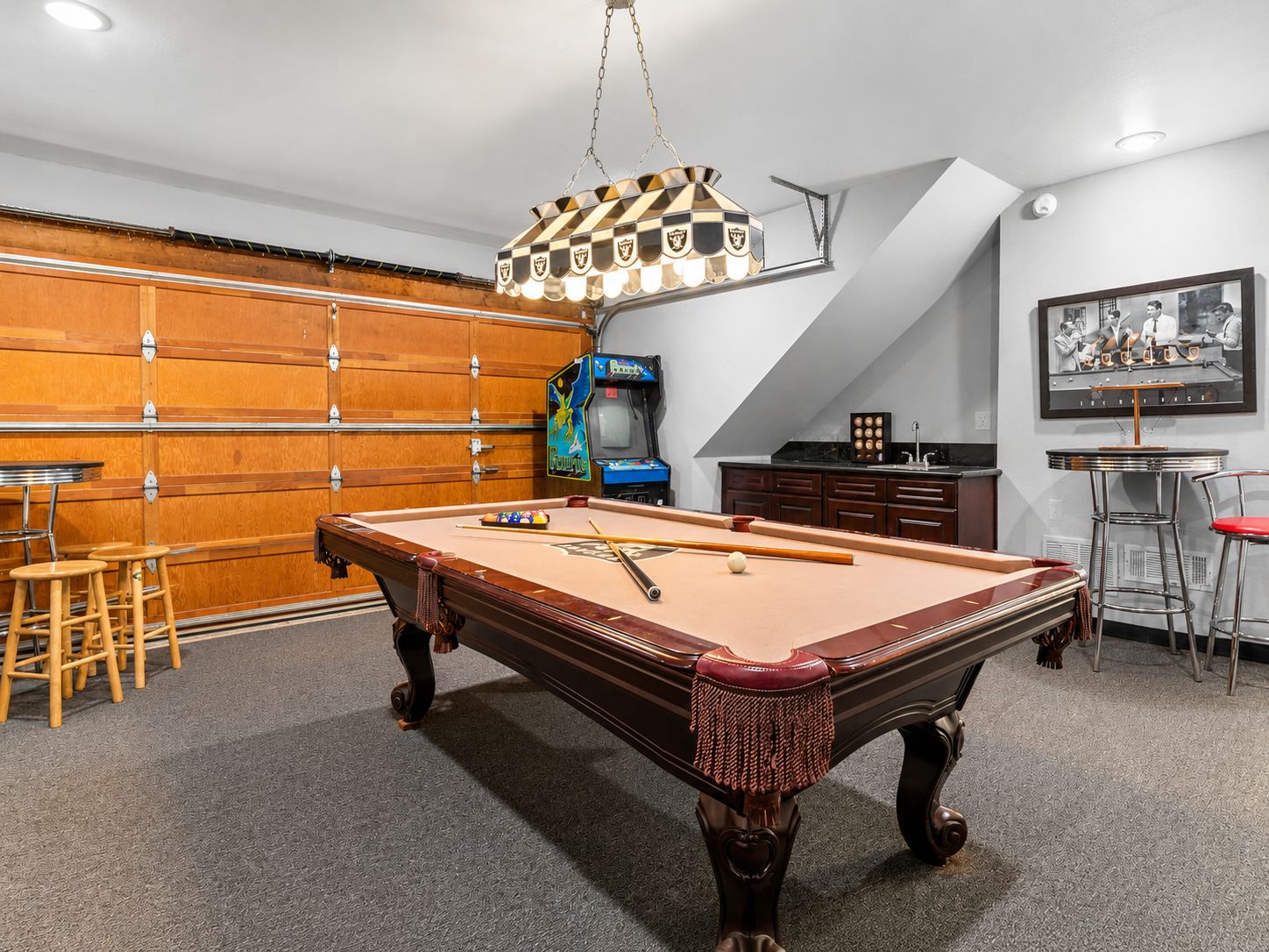 A pool table with stools, a bar area, and a gaming machine in a room with a sloped ceiling and wooden cabinets.