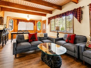 Een gezellige woonkamer met een houten plafond, een glazen salontafel en twee grijze banken met rode kussens.