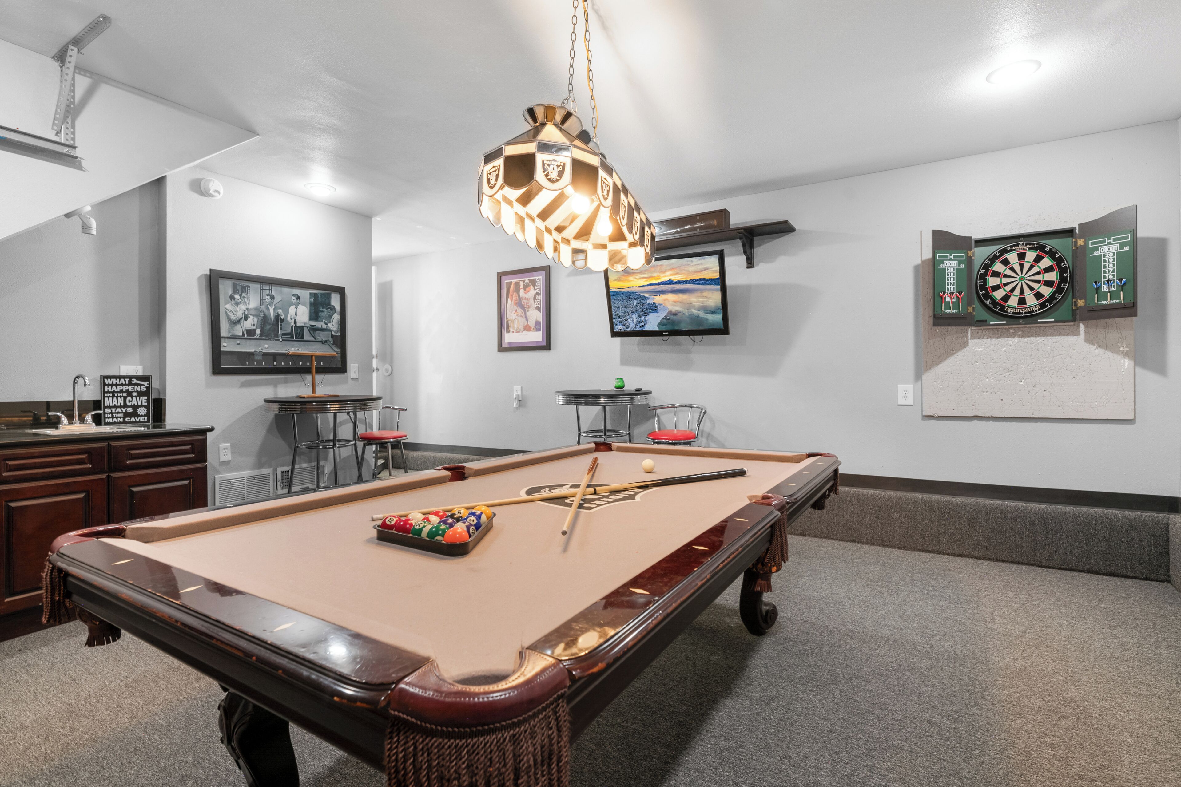 A pool table with billiard balls and cues, a dartboard, and a television mounted on the wall.
