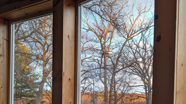 A room with a large window overlooking a lake and trees, a round table, and a patterned chair. - Apple River Retreat home in Amery  (Amery)