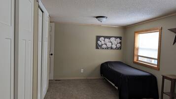 Une chambre avec un lit simple, une fenêtre avec des stores, un luminaire au plafond et un tableau accroché au mur.