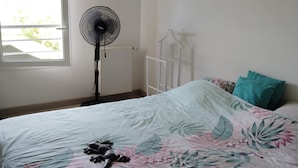 A bedroom with a bed, a standing fan, a radiator, and a window with a view of greenery.
