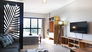 A modern living room with a sofa, a TV, a wooden entertainment unit, and a large window offering a view of the outdoors.